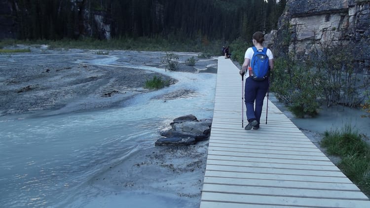 Lisa is walking away from the camera wearing hiking clothes and holding two walking sticks, she's crossing a wooden slatted bridge over salt plains