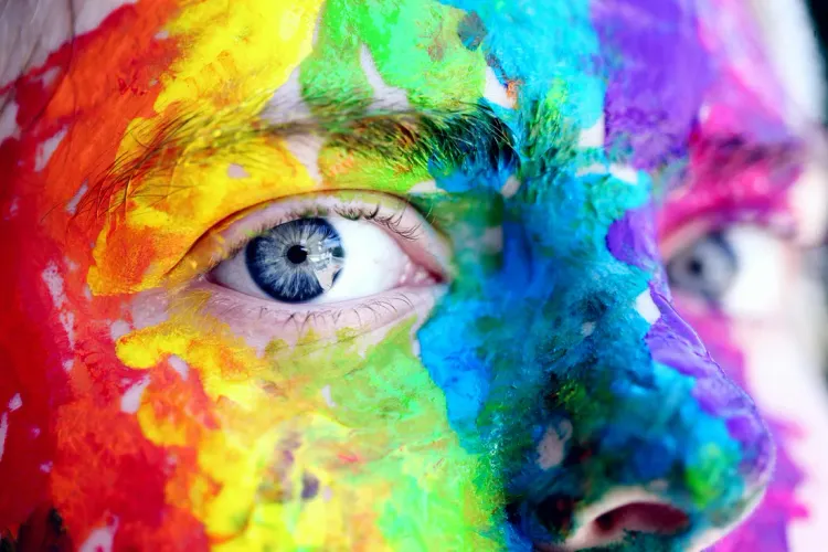 The eyes and nose of a Caucasian face with one blue eye visible. The face is covered in red, yellow, green, blue, purple and pink paint.