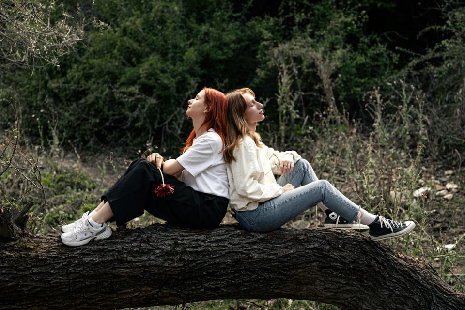 Two women sitting back to back on a fallen log in a forest. Both have their eyes closed and are holding a single stem flower.