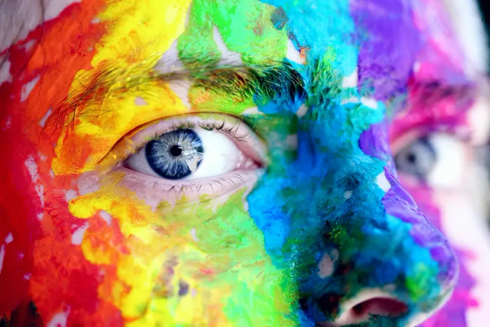 The eyes and nose of a Caucasian face with one blue eye visible. The face is covered in red, yellow, green, blue, purple and pink paint.