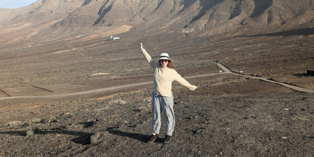 Lisa standing in front of a mountain range wearing a beige jumper and stripy trousers, sunglasses and a beige sunhat. She has her arms spread wide open as if gesturing to the mountains