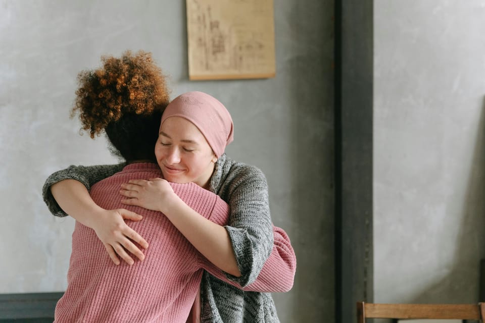 Two women are embracing in a grey walled room. The face of the woman we can see is smiling.